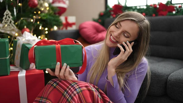 年轻的金发女人坐在家里的圣诞树旁 用智能手机聊天 — 图库照片