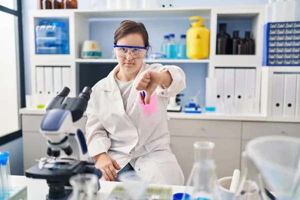 Hispanic girl with down syndrome working at scientist laboratory looking unhappy and angry showing rejection and negative with thumbs down gesture. bad expression.
