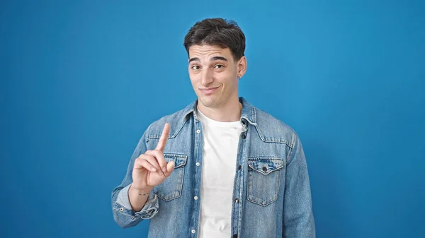 Young hispanic man standing with serious expression saying no with finger over isolated blue background