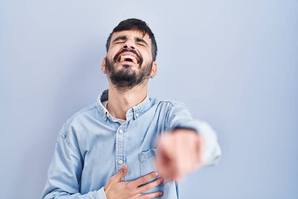 Young hispanic man with beard standing over blue background laughing at you, pointing finger to the camera with hand over body, shame expression 