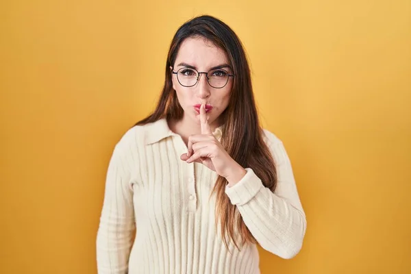 若いヒスパニック系の女性は黄色の背景の上に唇に指で静かにするように求めて立っている 沈黙と秘密の概念 — ストック写真