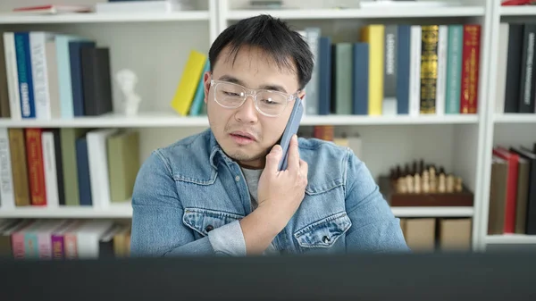 Young chinese man student using computer talking on smartphone at library university