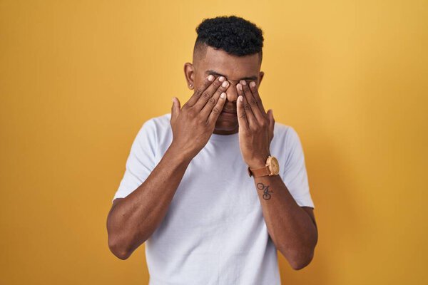 Young hispanic man standing over yellow background rubbing eyes for fatigue and headache, sleepy and tired expression. vision problem 