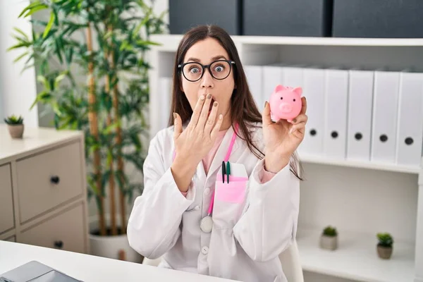 stock image Young doctor woman holding piggy bank covering mouth with hand, shocked and afraid for mistake. surprised expression 