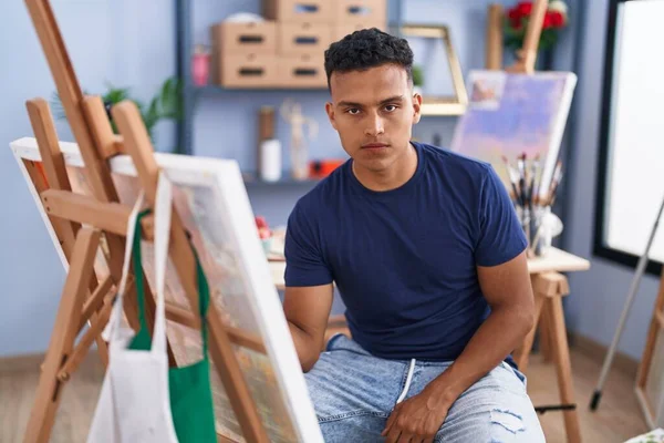 Young hispanic man painting sitting at art studio thinking attitude and sober expression looking self confident