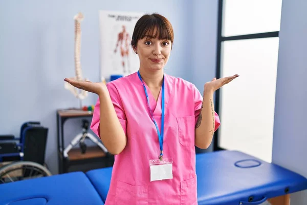 リハビリテーションクリニックで働く若いブルネットの女性は 腕や手を上げて混乱した表情 疑わしい概念 — ストック写真