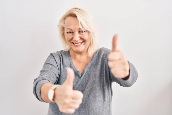 白人の背景の上に立つ中年の白人女性は 手で積極的なジェスチャーを行うことを承認します 親指アップ笑顔と成功のために幸せ 勝者のジェスチャー — ストック写真