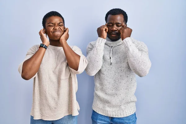 若いアフリカ系アメリカ人のカップルが青い背景の上に立ち 大きな音楽のノイズに悩まされた表情で耳を覆う 聴覚障害の概念 — ストック写真