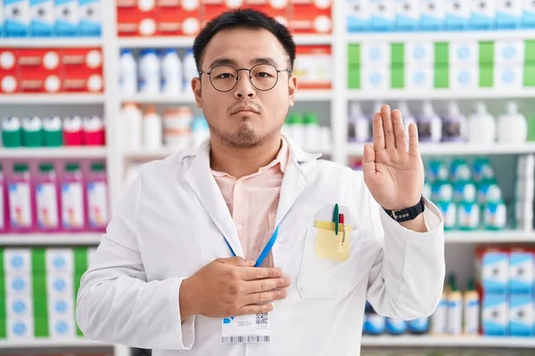 Chinese young man working at pharmacy drugstore swearing with hand on chest and open palm, making a loyalty promise oath