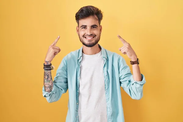 Молодий Іспаномовний Чоловік Татуюваннями Стоять Над Жовтим Тлом Посміхаючись Вказуючи — стокове фото