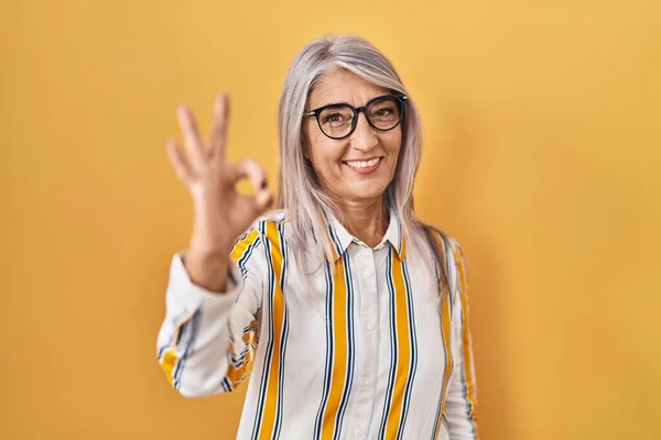 黄色い背景の上に白髪の中年女性が 手や指でOkサインをして積極的に笑顔で眼鏡をかけている 成功した表現 — ストック写真