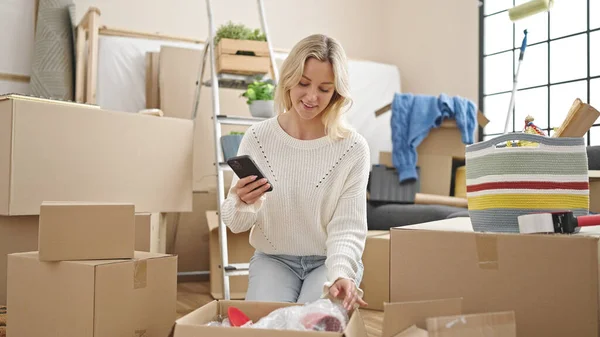 Young blonde woman unpacking cardboard box using smartphone at new home