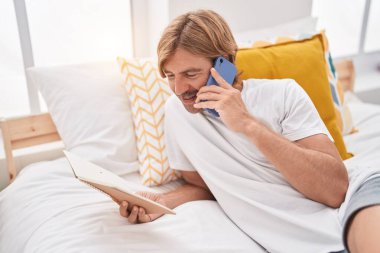Genç sarışın adam yatak odasında akıllı telefondan kitap okuyor.