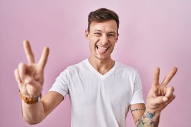 Pembe arka planda dikilen beyaz adam iki elini de zafer işareti yaparak parmaklarını göstererek gülümsüyor. İki numara.. 