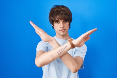 Latin kökenli genç adam mavi arka planda duruyor. Reddedilme ifadesi, kollarını kavuşturma, negatif işaret, kızgın surat. 