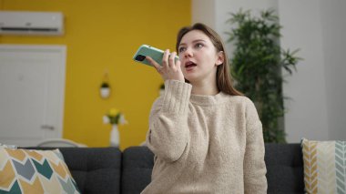 Sarışın genç kadın evdeki koltukta oturan akıllı telefonuyla sesli mesaj yolluyor.