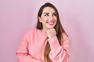 Young hispanic woman standing over pink background with hand on chin thinking about question, pensive expression. smiling and thoughtful face. doubt concept. 