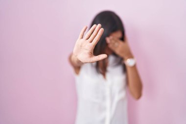 Esmer, pembe arka planda duran gözlüklü, elleriyle gözleri kapayan üzgün ve korku ifadeleriyle jest yapan genç bir kadın. utangaç ve olumsuz kavram. 