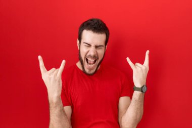Latin asıllı genç adam, gündelik kırmızı tişört giyip çılgın bir ifadeyle bağırıyor ve elleri havada rock sembolü yapıyor. Müzik yıldızı. ağır müzik konsepti. 