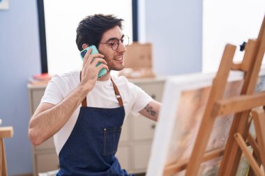 Young hispanic man artist talking on smartphone drawing at art studio