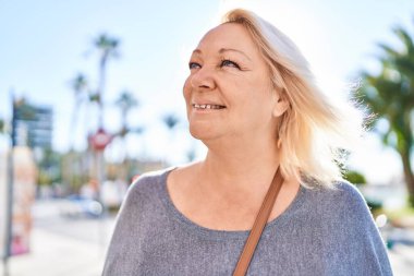 Orta yaşlı sarışın kadın sokakta kendinden emin bir şekilde gülümsüyor.