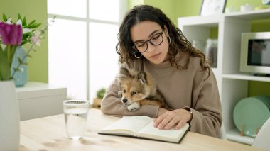 Köpekli genç İspanyol kadın yemek odasında masa başında kitap okuyor.