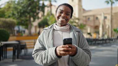 Parkta gülümseyen akıllı telefon kullanan Afrikalı Amerikalı kadın.