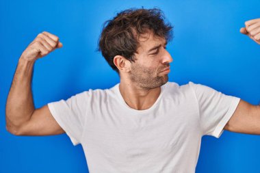 Mavi arka planda duran İspanyol genç adam gururla gülümseyen kol kaslarını gösteriyor. fitness konsepti. 