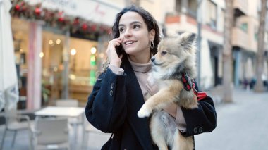 Köpeğiyle gülümseyen Latin kökenli genç bir kadın sokakta akıllı telefondan konuşuyor.