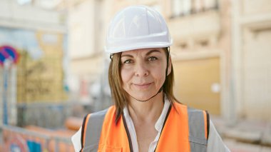 Orta yaşlı İspanyol kadın müteahhit inşaat alanında kendine gülerek duruyor.