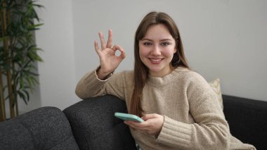 Young blonde woman using smartphone doing ok gesture at home