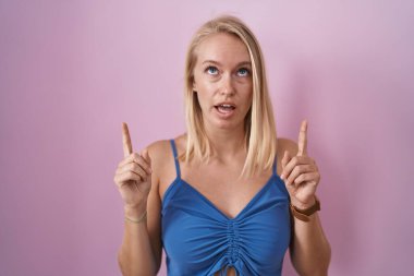 Young caucasian woman standing over pink background amazed and surprised looking up and pointing with fingers and raised arms. 