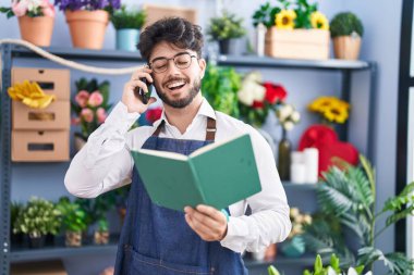 Young hispanic man florist talking on smartphone reading book at florist shop