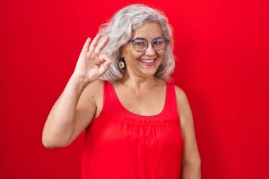 Kırmızı arka planda gri saçlı orta yaşlı bir kadın gülümsüyor ve el ve parmaklarıyla 