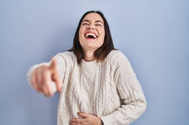 Genç esmer kadın mavi arka planda dikilip sana gülüyor, eli vücudunun üzerinde kamerayı işaret ediyor, utanç ifadesi. 