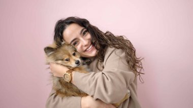 Köpeğiyle gülümseyen genç İspanyol kadın pembe arka planda duruyor.