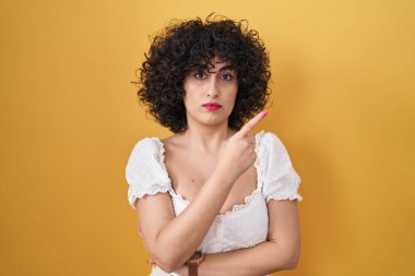Sarı arka planda duran kıvırcık saçlı, genç esmer kadın el parmağı yanda, ciddi ve sakin bir yüz ifadesi sergiliyor. 
