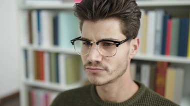 Young hispanic man student sitting on table with relaxed expression at library university