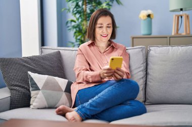 Evdeki koltukta oturan orta yaşlı bir kadın akıllı telefon kullanıyor.