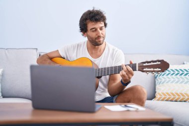 Latin kökenli genç bir adam evdeki kanepede oturup online gitar dersi alıyor.