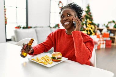 African american woman eating hamburger talking on smartphone sitting on table by christmas tree at home