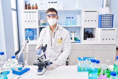 Laboratuarda mikroskop kullanan genç İspanyol bilim adamı tıbbi maske takıyor.