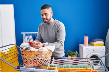 Young latin man talking on smartphone hanging clothes on clothesline at laundry room