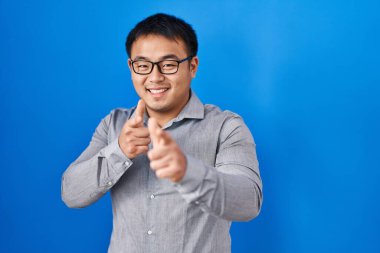 Mavi arka planda duran genç Çinli adam mutlu ve komik yüzlü parmaklarını kameraya doğrultuyor. iyi enerji ve titreşimler. 
