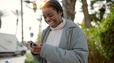 Parkta gülümseyen akıllı telefon kullanan Afrikalı Amerikalı kadın.