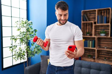 Young hispanic man training using dumbbell sitting on sofa at home