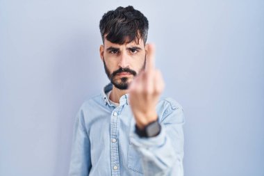 Young hispanic man with beard standing over blue background showing middle finger, impolite and rude fuck off expression 