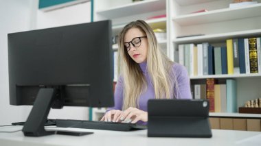 Young blonde woman student using computer and touchpad studying at library university