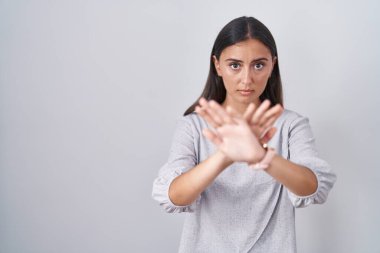 Beyaz arka planda duran Latin kökenli genç bir kadın. Reddedilme ifadesi. Kollarını kavuşturup, avuç içleri negatif işaretlerle dolu. 