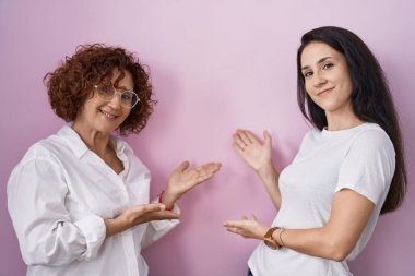 Hispanic mother and daughter wearing casual white t shirt over pink background inviting to enter smiling natural with open hand 